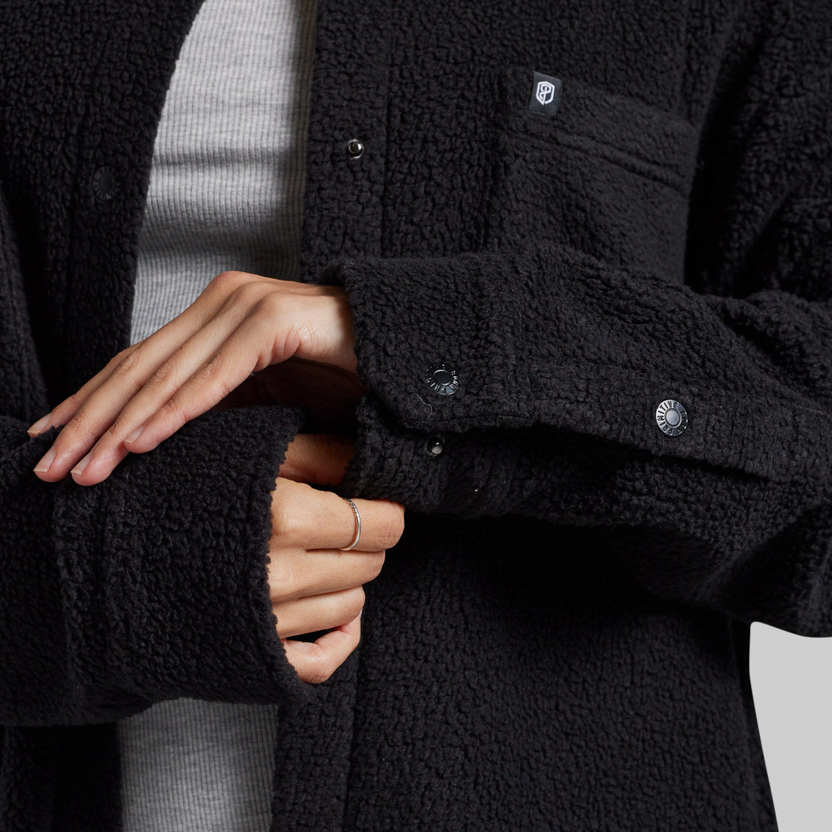 Close-up of a person wearing a black sherpa fleece button-front jacket over a light gray ribbed top, with both hands visible adjusting the sleeve cuff.