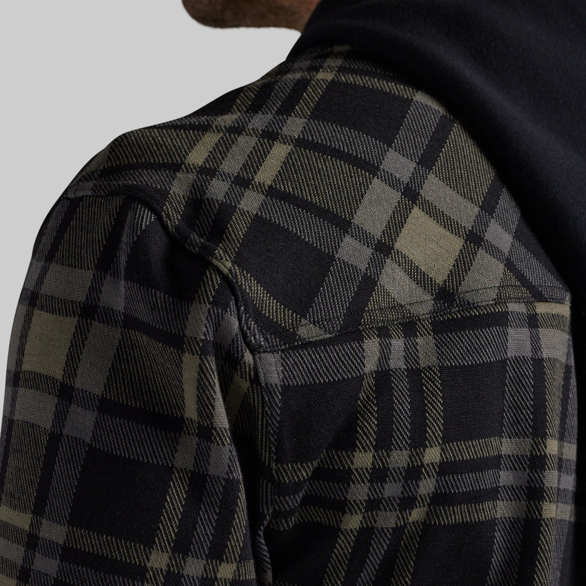 Close-up of a person wearing a dark plaid overshirt or jacket with a black hood, showing the shoulder and upper chest against a light gray background.
