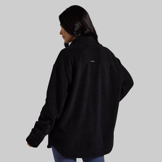 A woman shown from the back wearing an oversized black fleece jacket against a solid light gray background.