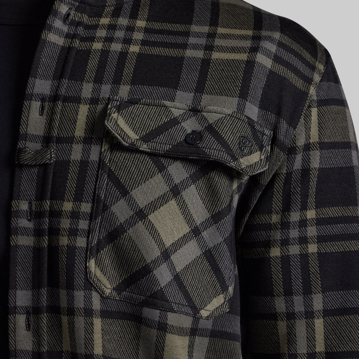 Close-up of a black and greu plaid flannel shirt, showing a chest pocket with a button flap and a small shield logo patch.