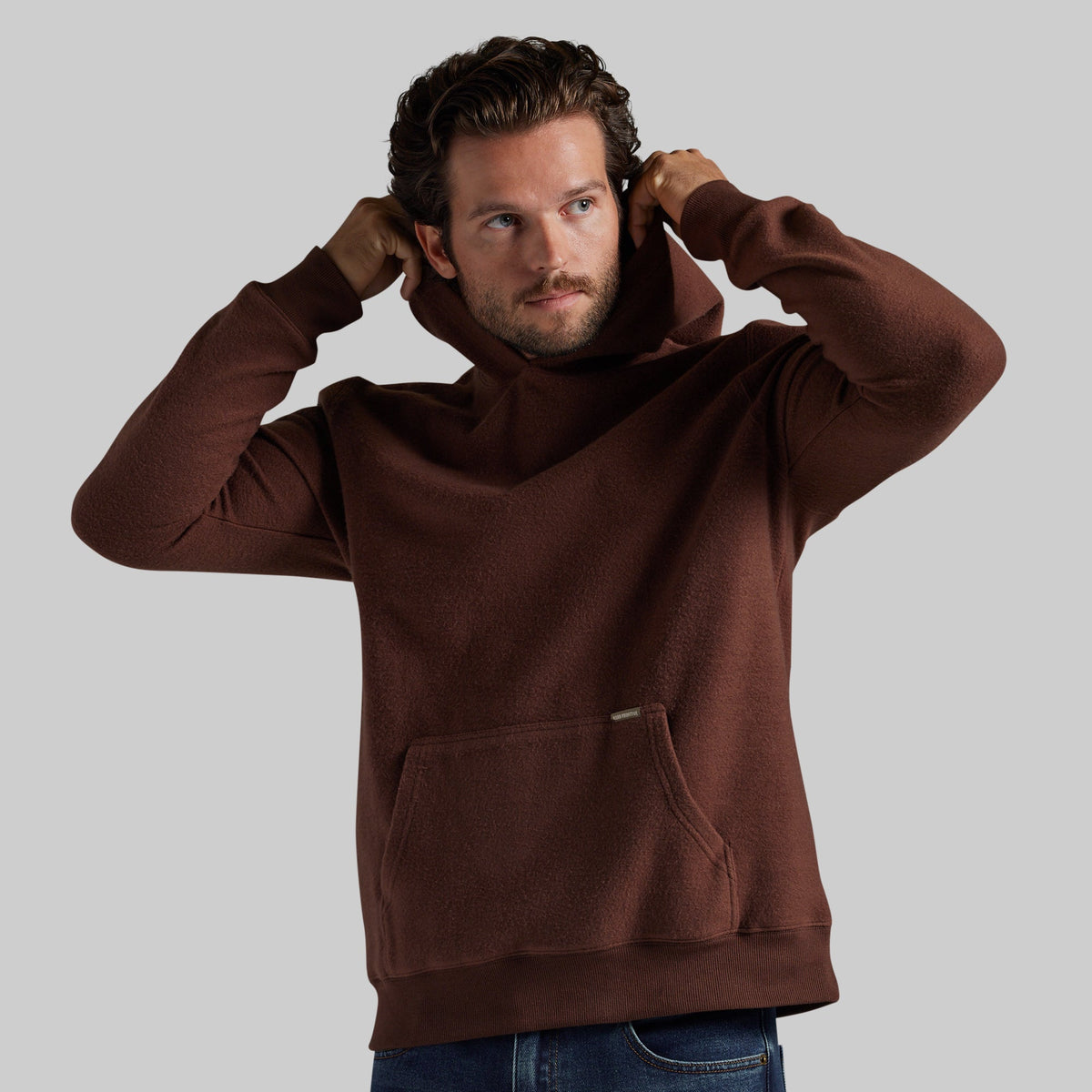 A man wearing a brown fleece pullover hoodie with a kangaroo pocket, shown from the waist up against a solid gray background.