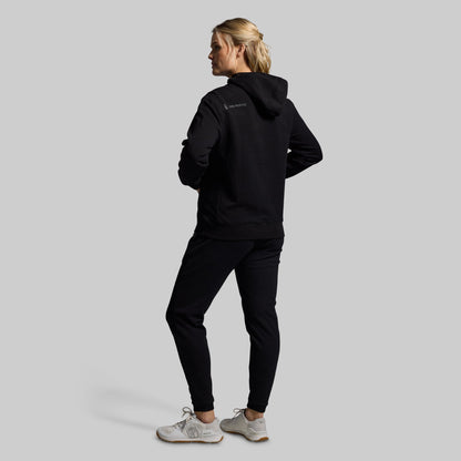 Person wearing a black hoodie and pants with a visible brand logo on a light gray background