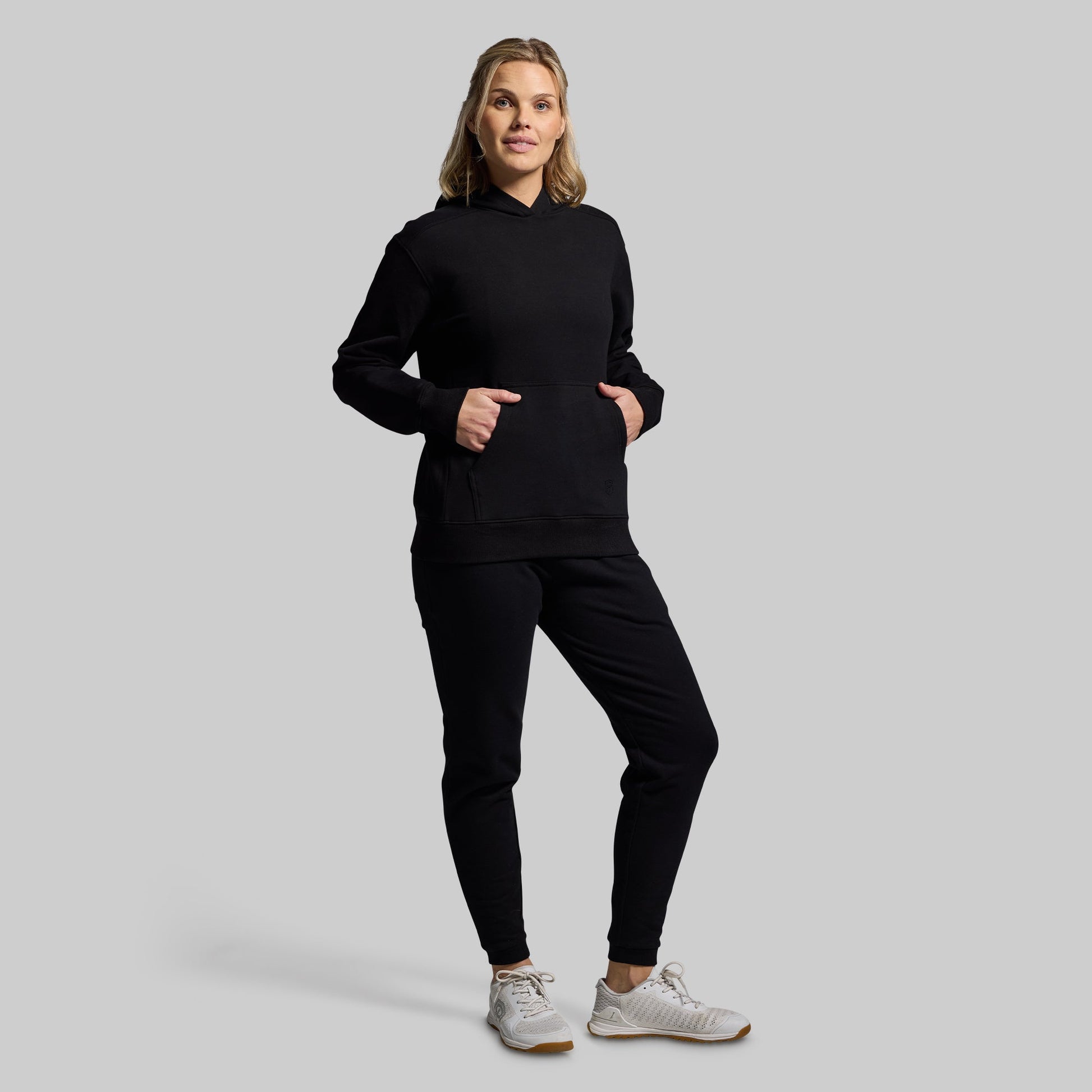 A woman modeling a black pullover hoodie and matching black jogger pants against a solid light gray studio background.