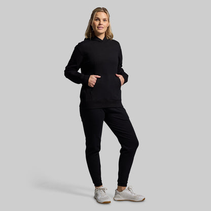A woman modeling a black pullover hoodie and matching black jogger pants against a solid light gray studio background.