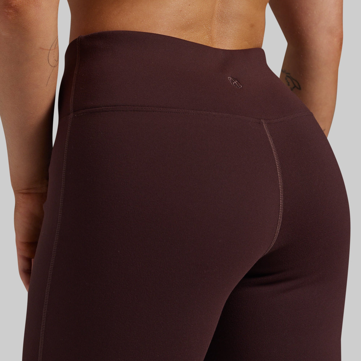 Close-up back view of a person wearing high-waisted leggings in a deep plum-brown color against a light gray studio background.