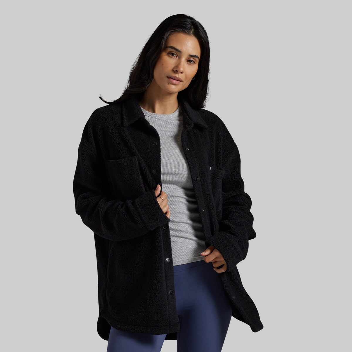 A woman modeling a black sherpa fleece button-up overshirt over a gray t-shirt and blue leggings, set against a solid light gray studio background.