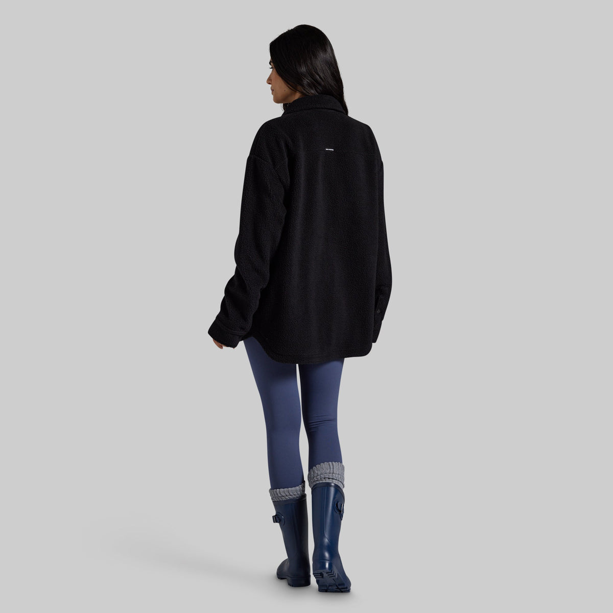 A woman is shown from the back wearing a black fleece jacket, blue leggings, and dark blue rain boots against a solid gray studio background.