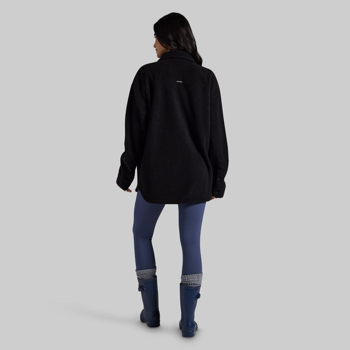 A woman is shown from behind wearing a black fleece pullover, blue leggings, and blue rain boots against a solid gray studio background.