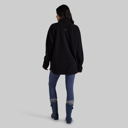 A woman is shown from behind wearing a black fleece pullover, blue leggings, and blue rain boots against a solid gray studio background.