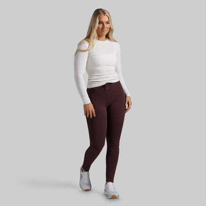 A woman modeling a white long-sleeve top and dark burgundy leggings with white sneakers, standing against a solid light gray background.