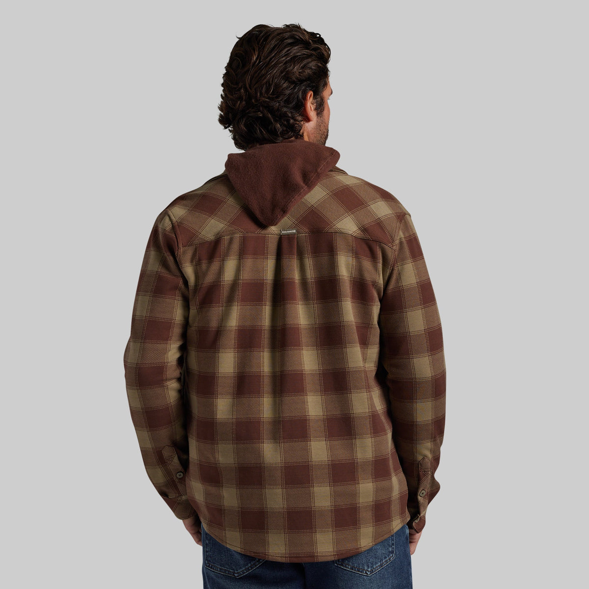 A man viewed from behind wearing a brown and tan plaid hooded flannel shirt jacket, paired with blue jeans, against a solid gray background.