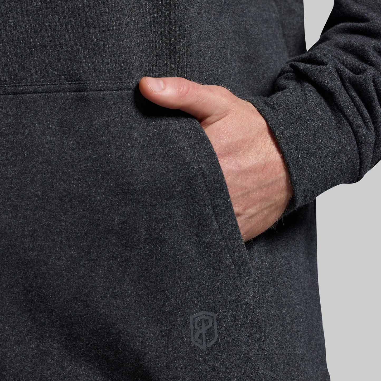 Person wearing a dark gray sweatshirt with a logo on the kangaroo pocket against a light gray background