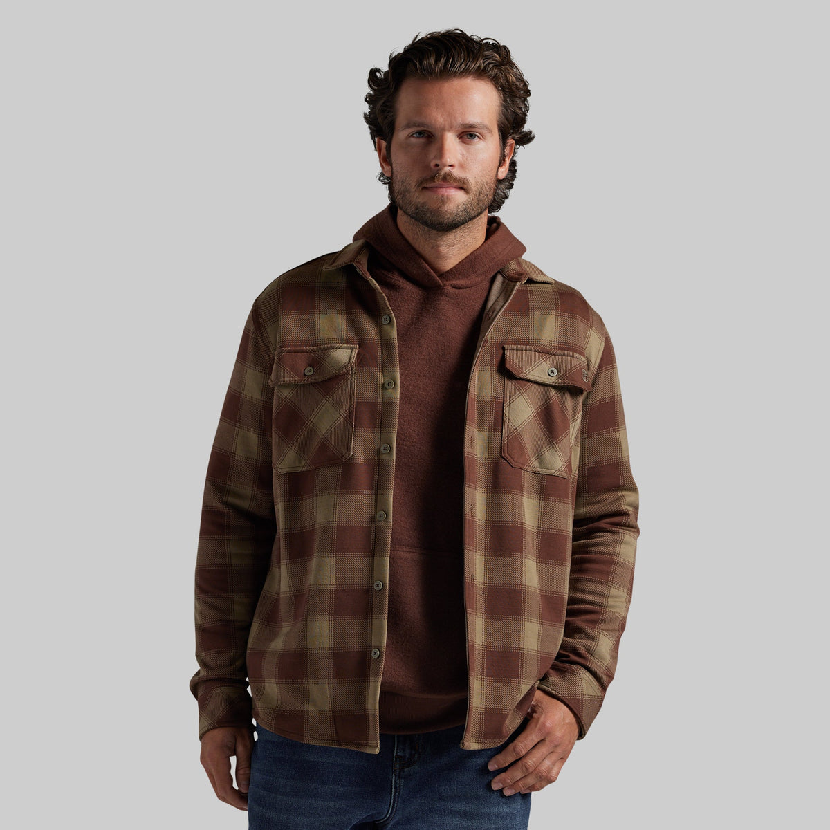 A man wearing a brown and tan plaid overshirt over a brown hoodie and blue jeans, standing against a solid gray background.