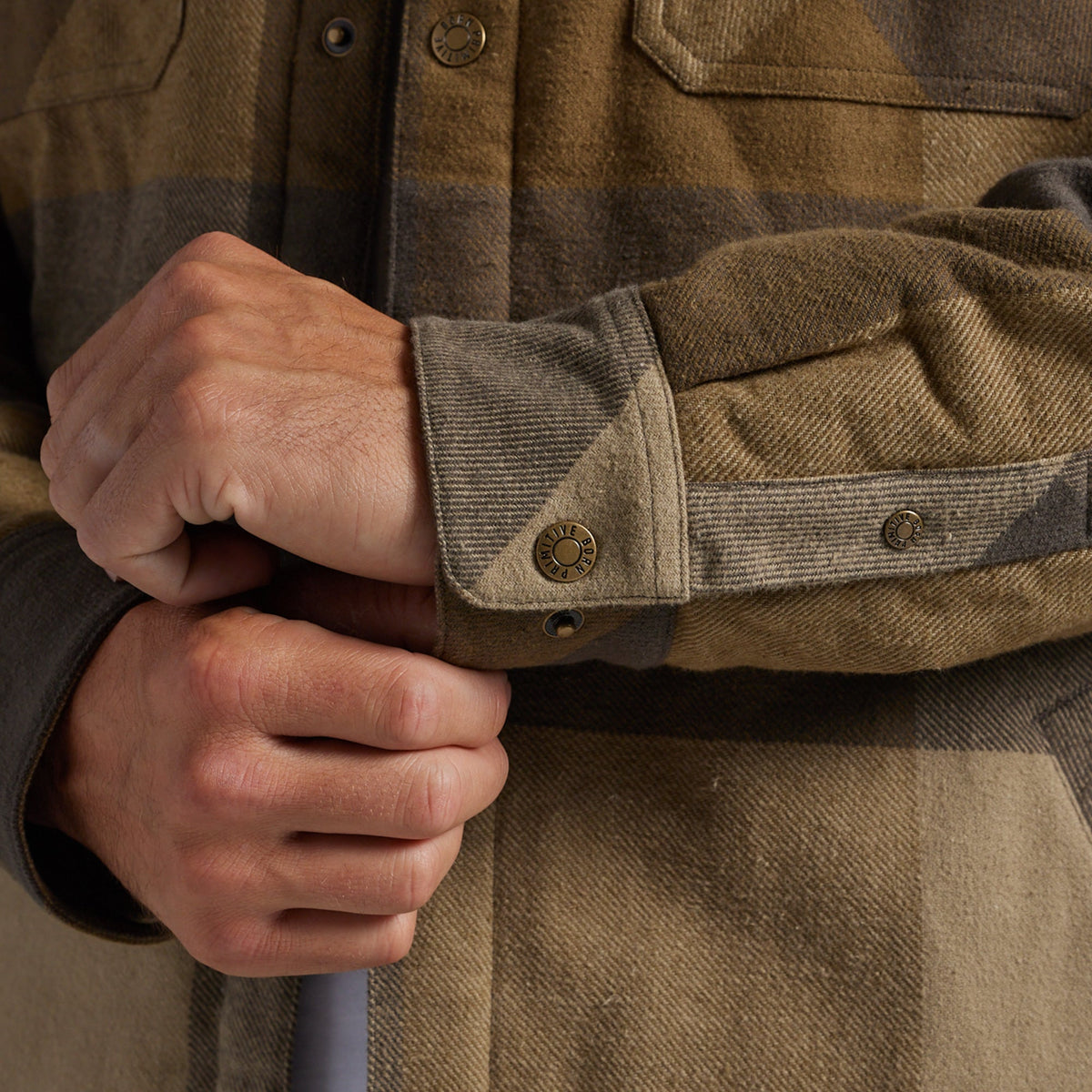 Close-up of a person's hands adjusting the cuff of a brown and tan plaid flannel jacket, showing the fabric and snap-button cuff.