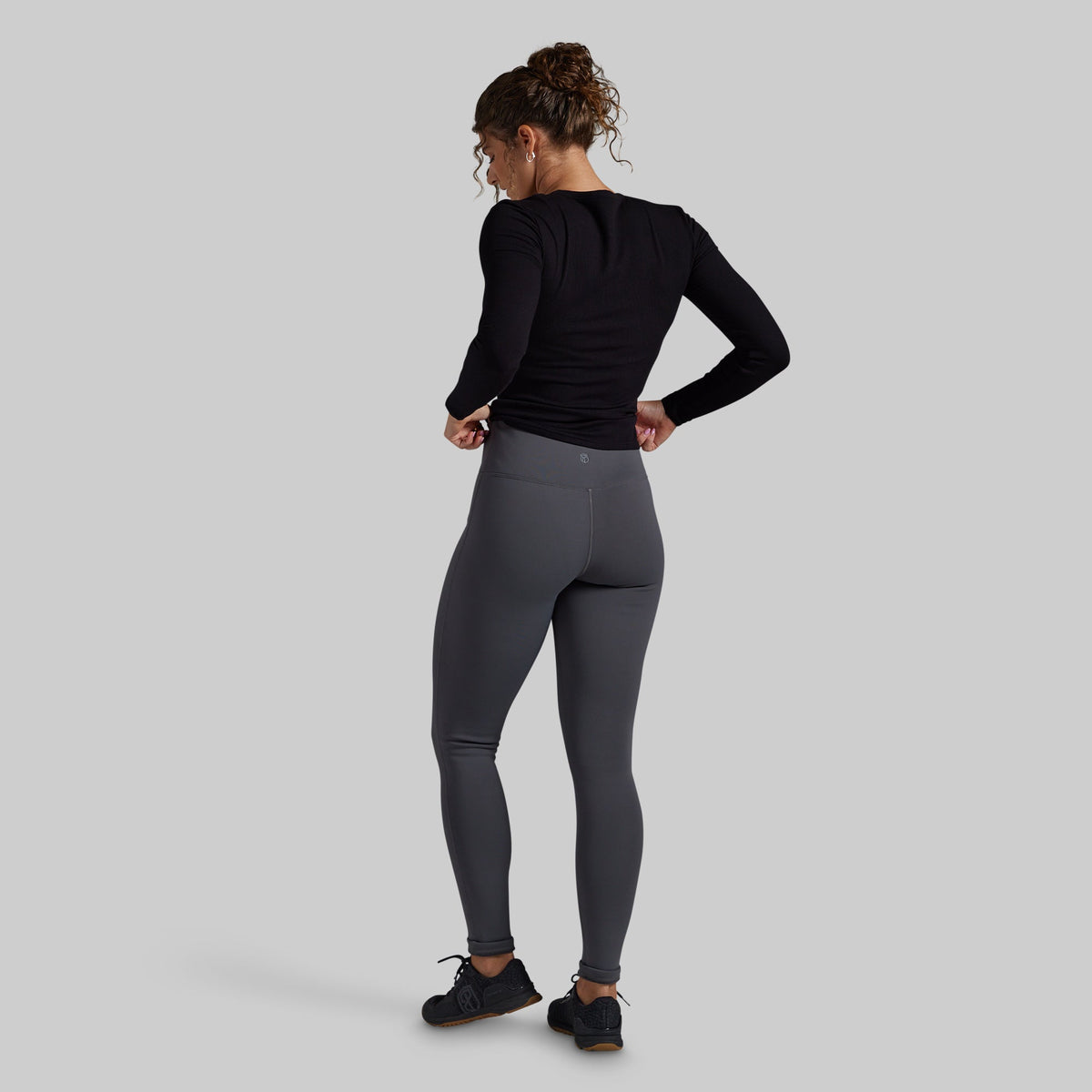 A woman shown from the back wearing dark gray high-waisted fleece lined leggings, a black long-sleeve top, and black sneakers against a solid gray studio background.