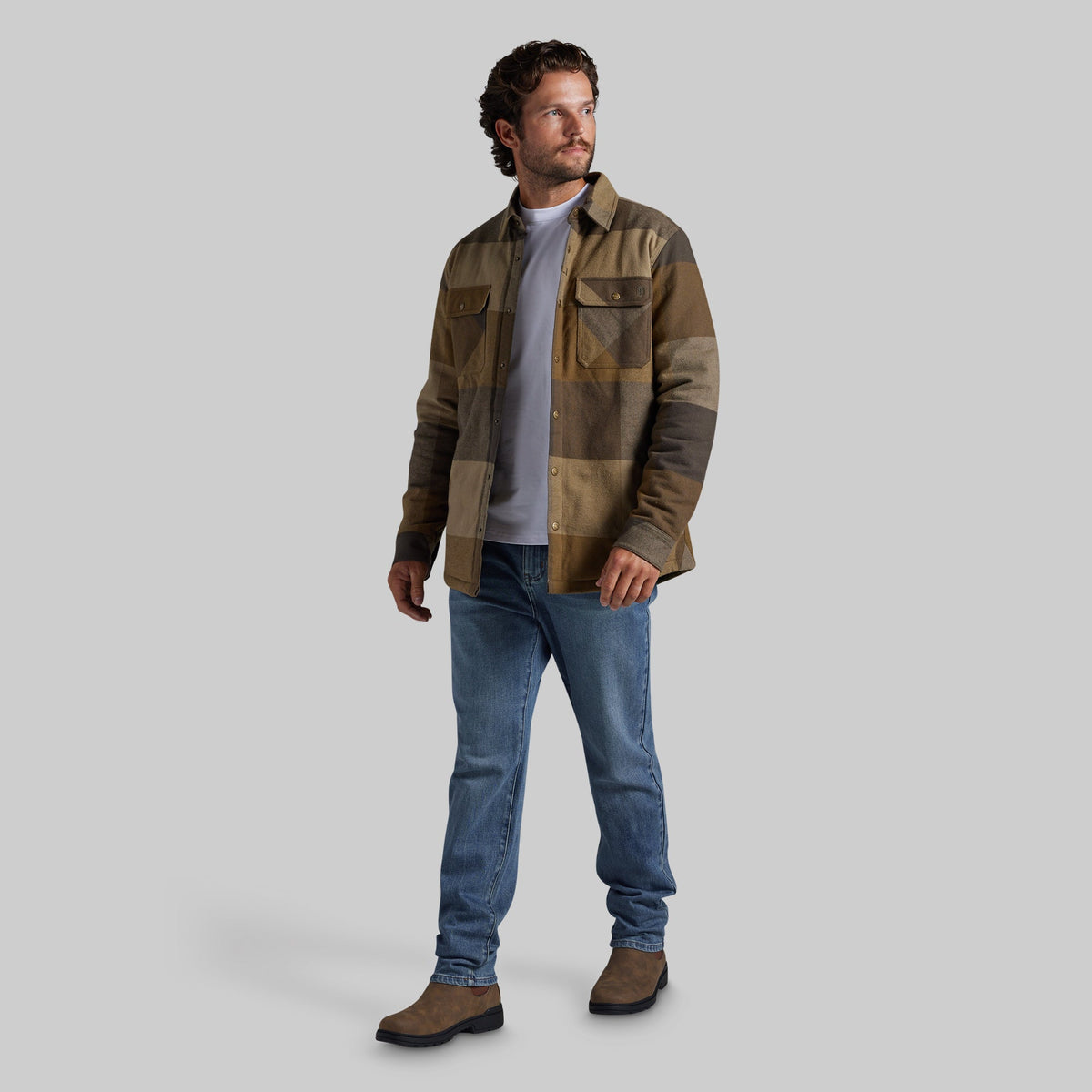 A man wearing a brown and tan patchwork flannel jacket, blue jeans, and brown boots, standing against a solid gray studio background.
