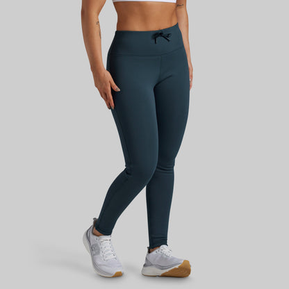 A model wearing high-waisted, full-length fleece lined leggings in a dark teal blue color, paired with white sneakers, shown from the waist down against a solid light gray background.