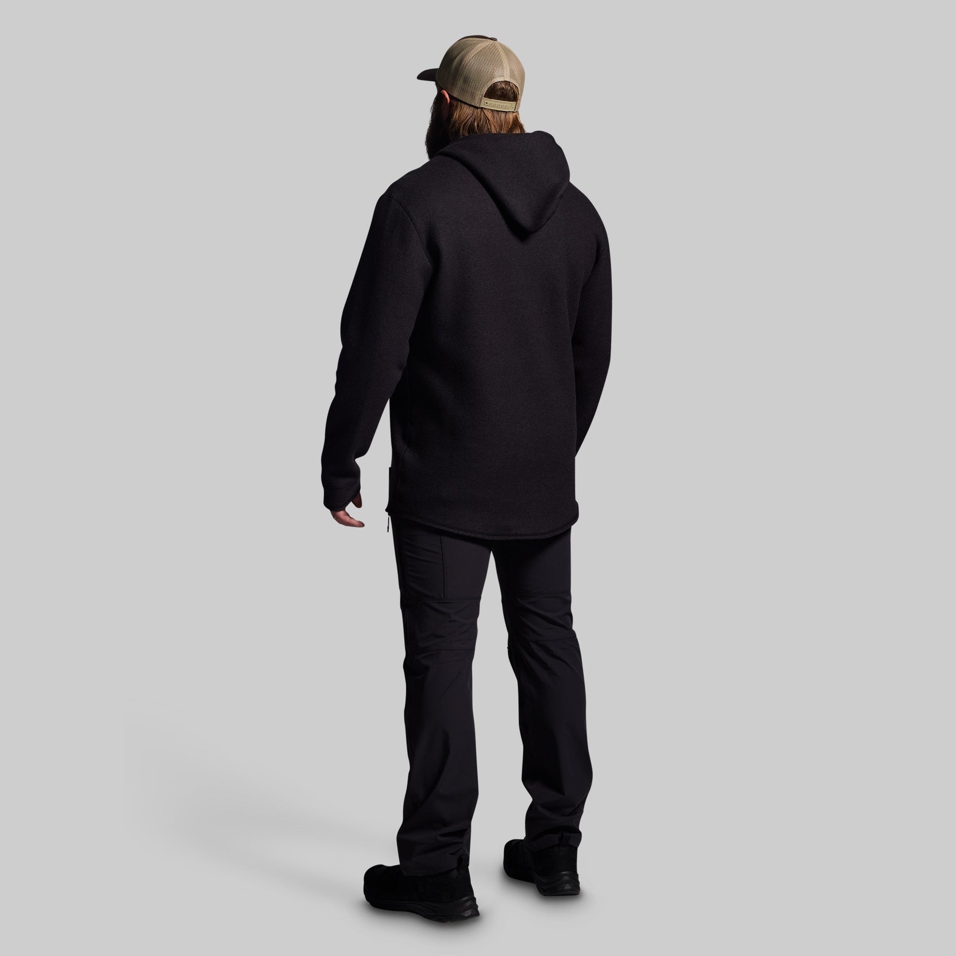 Person wearing a black hoodie and pants with a beige cap on a light gray background