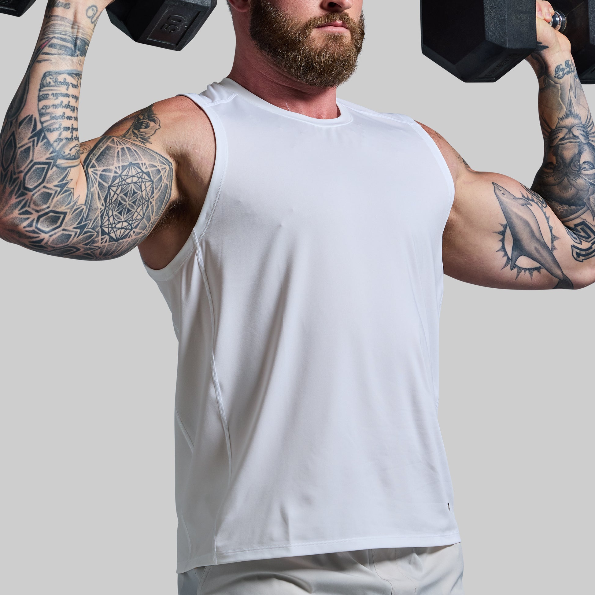 Man with tattoos lifting dumbbells wearing a white tank top on a plain background