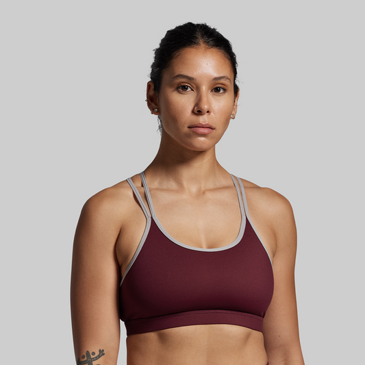 Woman wearing a maroon sports bra with gray straps against a plain background