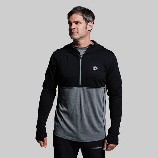 A man wearing a black and gray half zip hoodie with a hood, designed with a lighter gray panel on the lower half and a small logo on the chest.