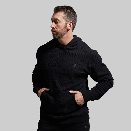 A man wearing a black hoodie with a waffle-textured material, standing with hands in pockets.