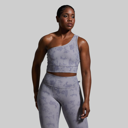 Woman wearing a gray one-shoulder sports bra and leggings against a plain background